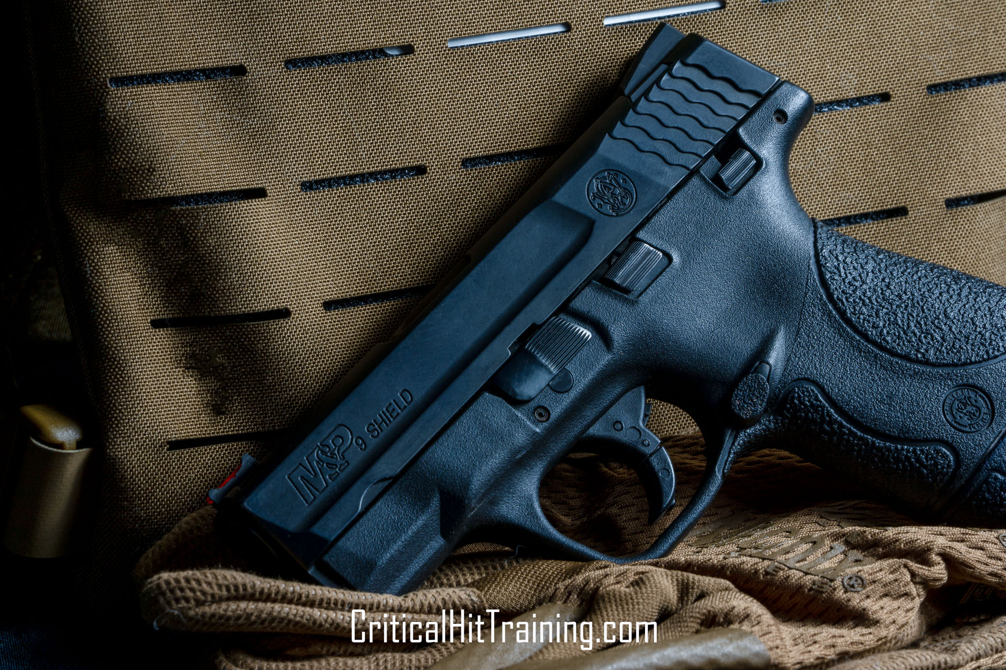 Image of an M&P Shield handgun positioned in front of a set of body armor, showcasing the firearm's compact design and the tactical gear's rugged details, set against a neutral background.